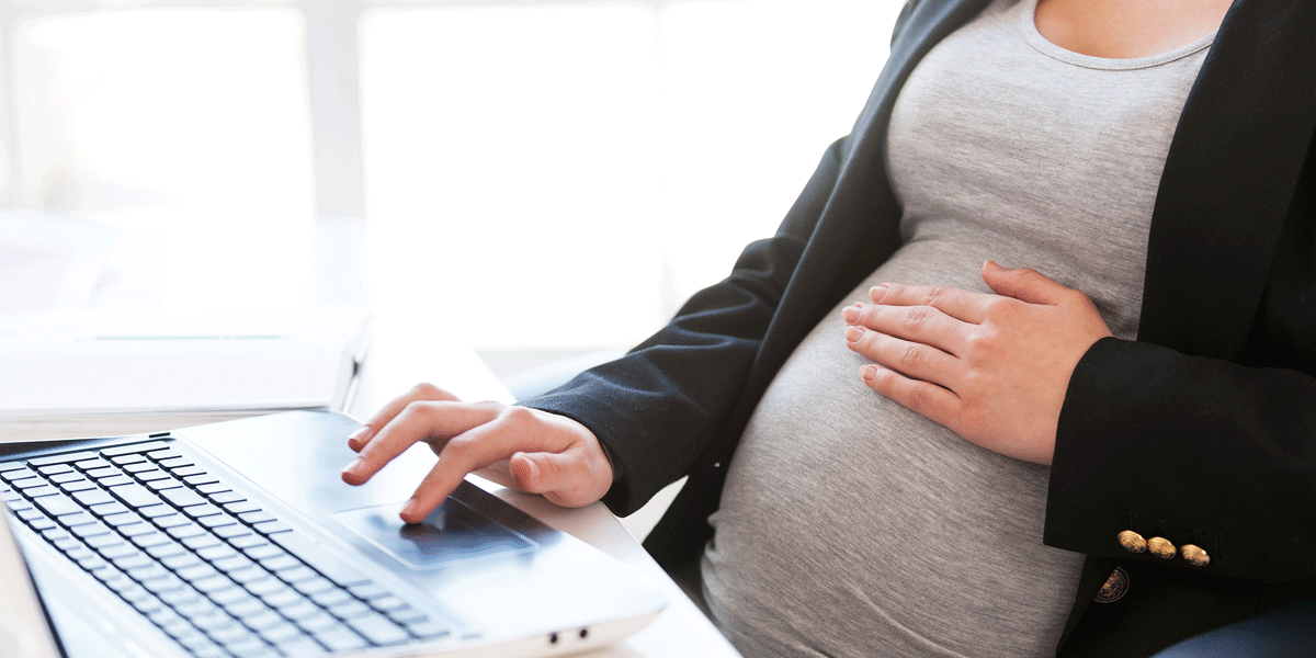 A close-up of a woman's pregnant belly. Her hand rests on her belly as she works on a laptop.